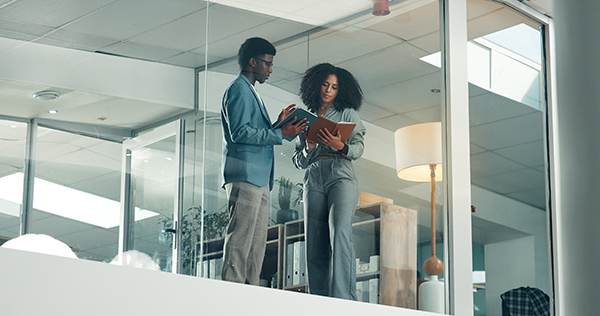 Two professionals standing in a glass office reviewing documents together.