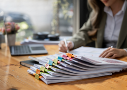 Stack of organized documents on a desk with a person reviewing paperwork in the background.