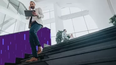 Person walking down a staircase in a modern office building while using a tablet and talking on a phone.