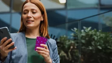 Person using a smartphone while holding a payment card outside a modern office building.