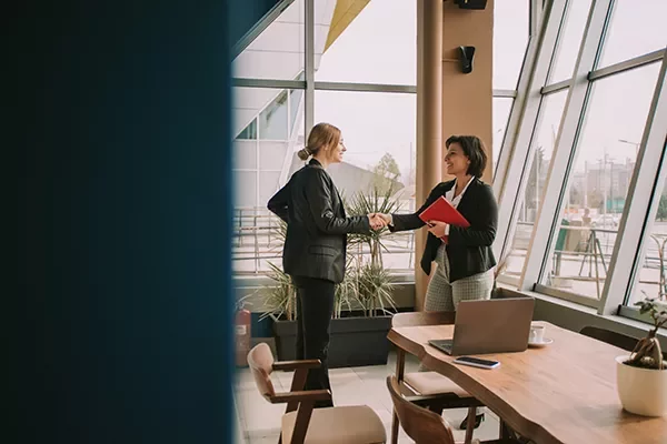 Two professionals shaking hands while holding documents in a modern office with large windows.