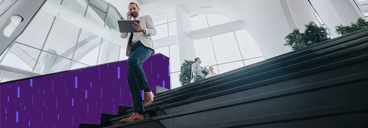 Person walking down a staircase in a modern office building while using a tablet and talking on a phone.