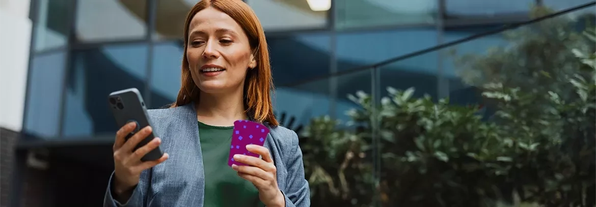 Person using a smartphone while holding a payment card outside a modern office building.